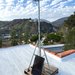 A completely set up sensor with a box at the top of a tall metal stand. Everything is mounted on a wooden pallet riser an sits atop a flat roof with trees and dramatic blue skies with white clouds in the background.