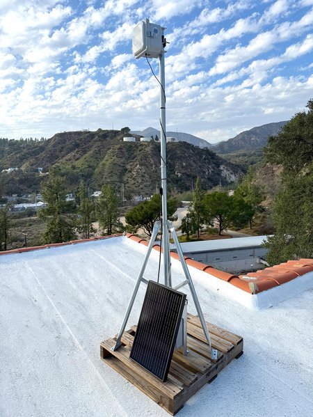 A completely set up sensor with a box at the top of a tall metal stand. Everything is mounted on a wooden pallet riser an sits atop a flat roof with trees and dramatic blue skies with white clouds in the background.