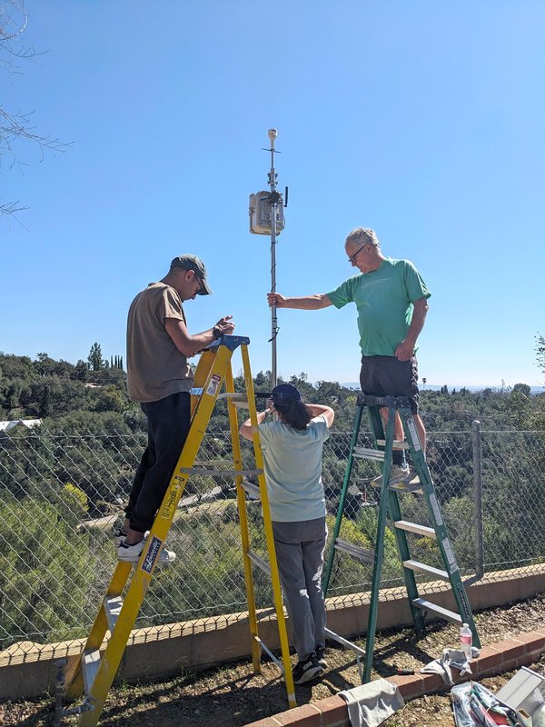 Two men stand on ladders and a woman stands on the ground, working to install a QuantAQ sensor such that it extends up from a fence.