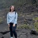 A person in black pants, a blue jacket, pink tank, and necklace stands on a dark gray ground of lava rock dotted with vigorous plants, with green trees rising on a steep hills in the background