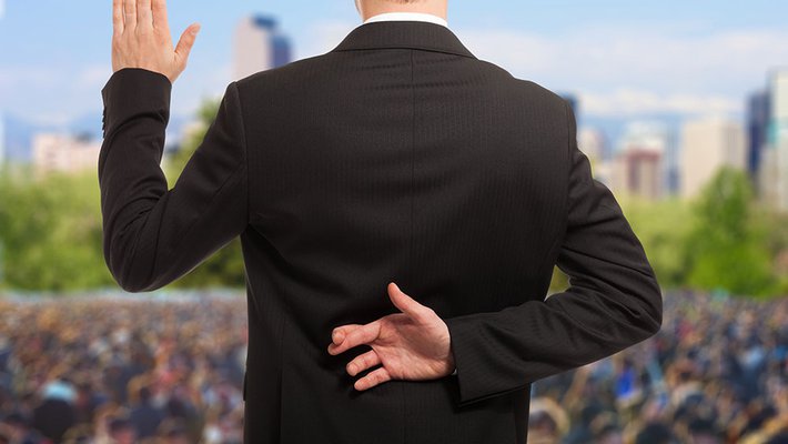 A politician stands on a stage in front of a large crowd taking an oath of office. His fingers are crossed behind his back 