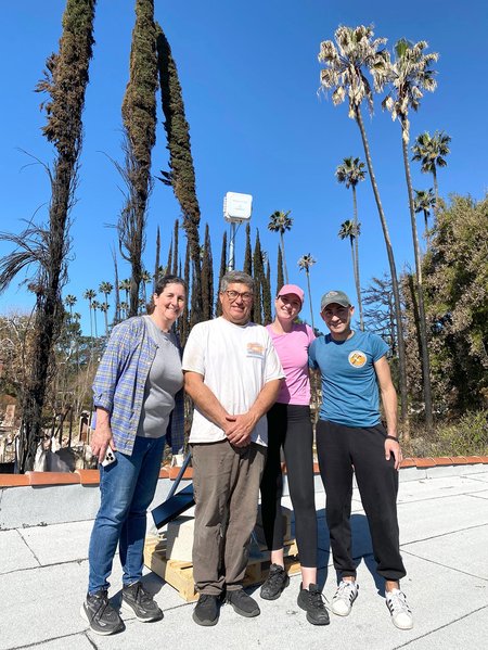 Two women and two men pose with an air quality monitoring sensor on a roof with burned trees in the background.