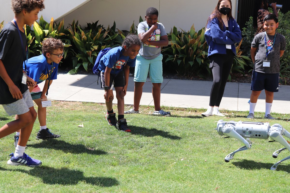 Children with one adult look at a doglike robot walking on grass.