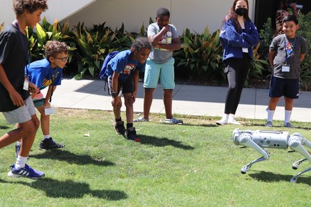 Children with one adult look at a doglike robot walking on grass.