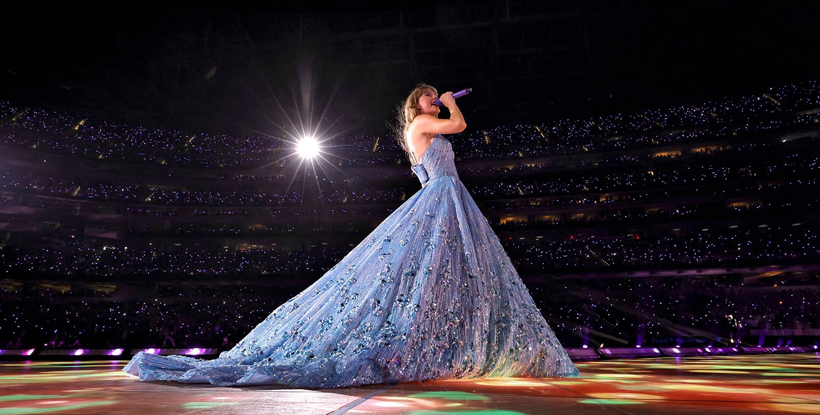 Taylor Swift, seen from the side, wearing a shimmering blue ball gown. In the background, you can see many tiny lights from the thousands of fans in SoFi Stadium.