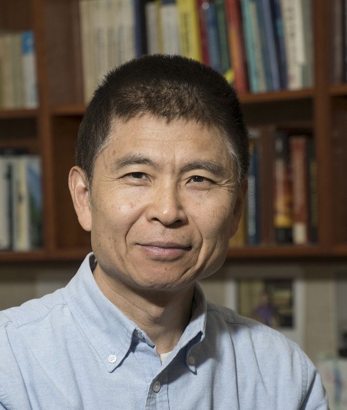 A portrait of Zhen-Gang Wang. He wears a dress shirt and appears in front of shelves of books.