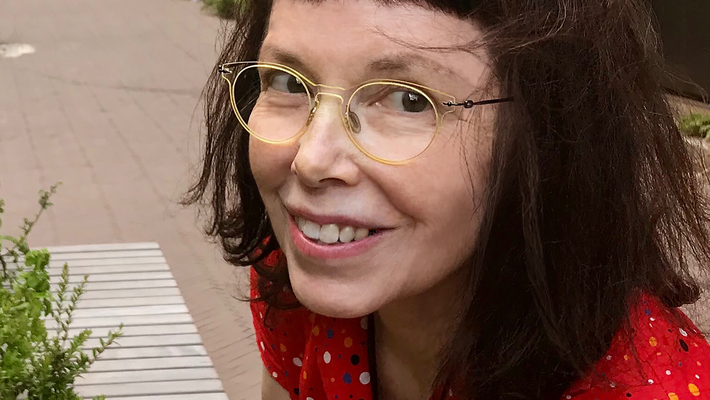 A portrait of artist Leslie Thornton. She wears a red dress and glasses.